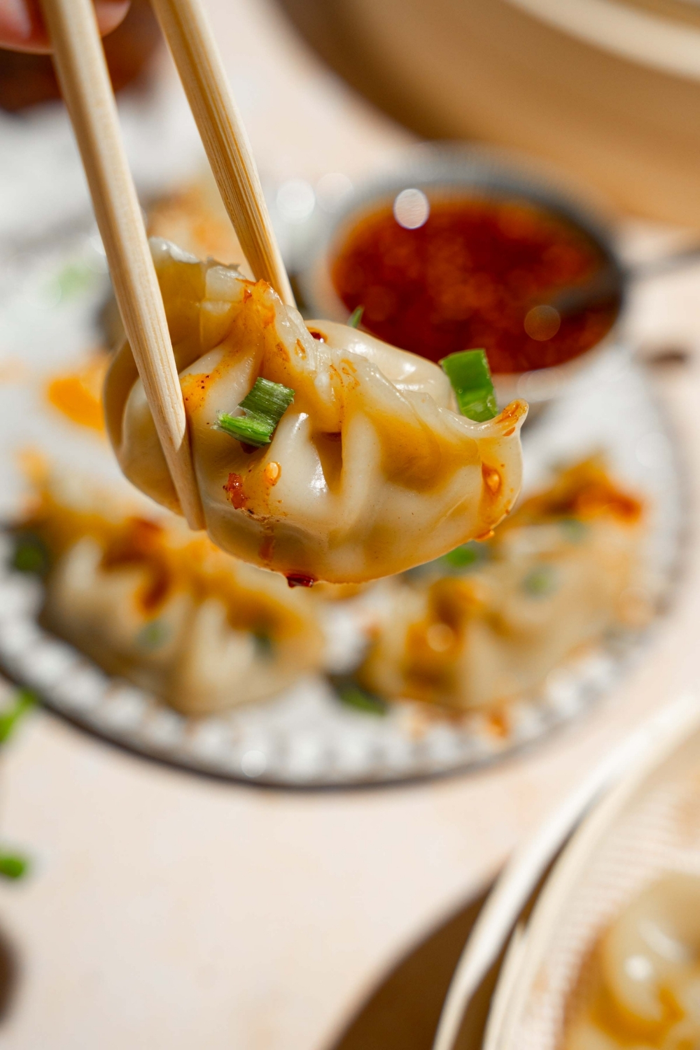 A close up of a pair of chopsticks holding a chicken dumpling garnished with sliced green onion. There is a plate of dumplings with a small bowl of chili oil sauce blurred in the background.