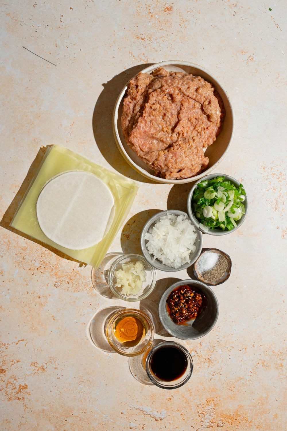 A tan counter with ingredients to make chicken dumplings including ground chicken, dumpling wrappers, green onion, onion, garlic, ginger, soy sauce, chili oil, and more.