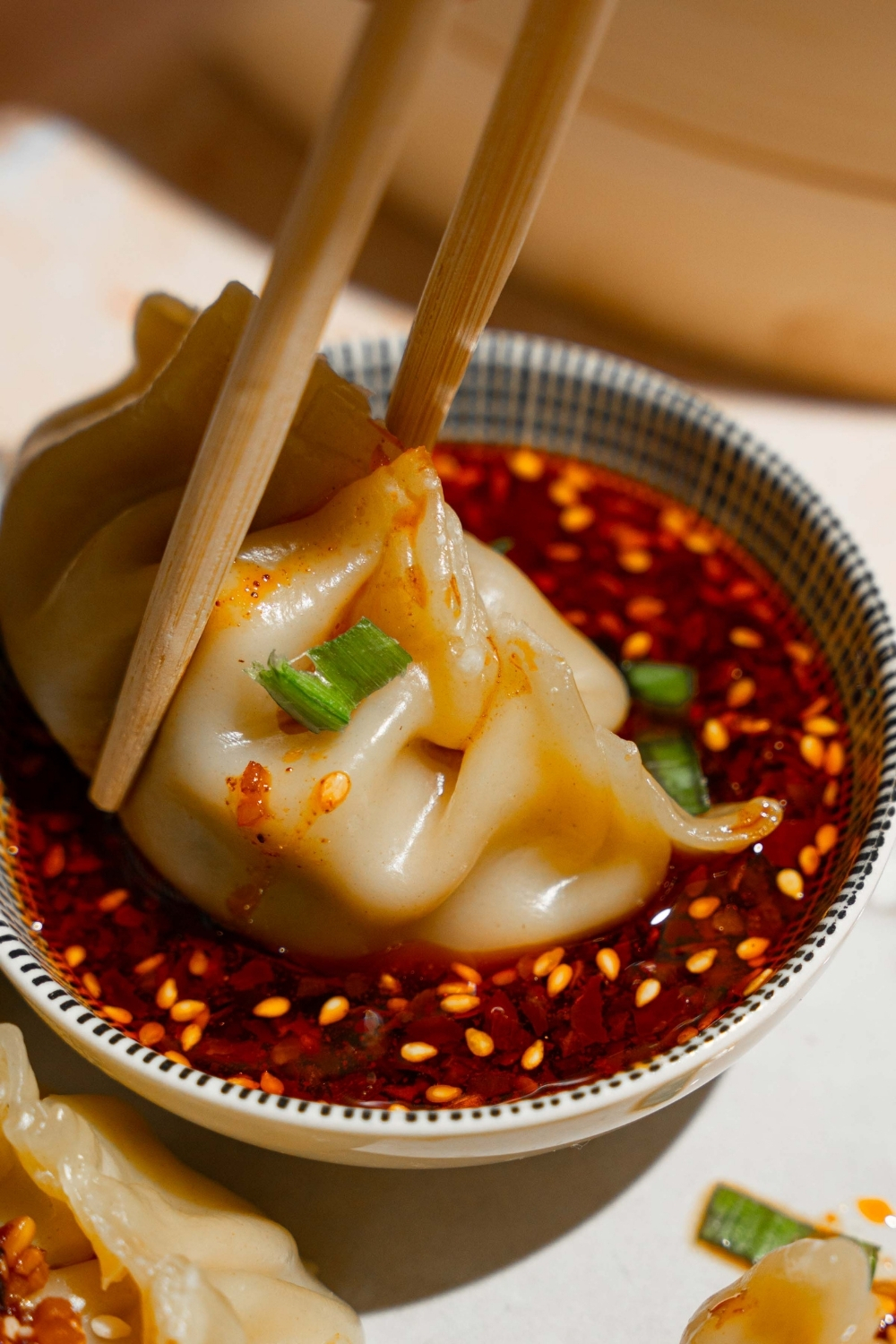 A pair of chopsticks dipping a chicken dumpling into a small bowl of chili oil sauce. The bowl is on a tan counter.