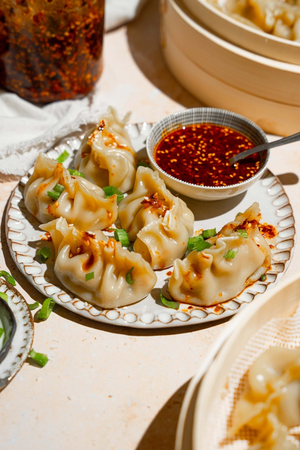 A plate with steamed chicken dumplings with a bowl of chili oil sauce. The plate is on a tan counter with a basket of steamed dumplings and white cloth napkin.