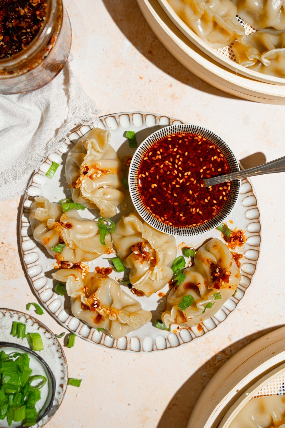 A plate with steamed chicken dumplings with a bowl of chili oil sauce. The plate is on a tan counter with a basket of steamed dumplings and white cloth napkin.