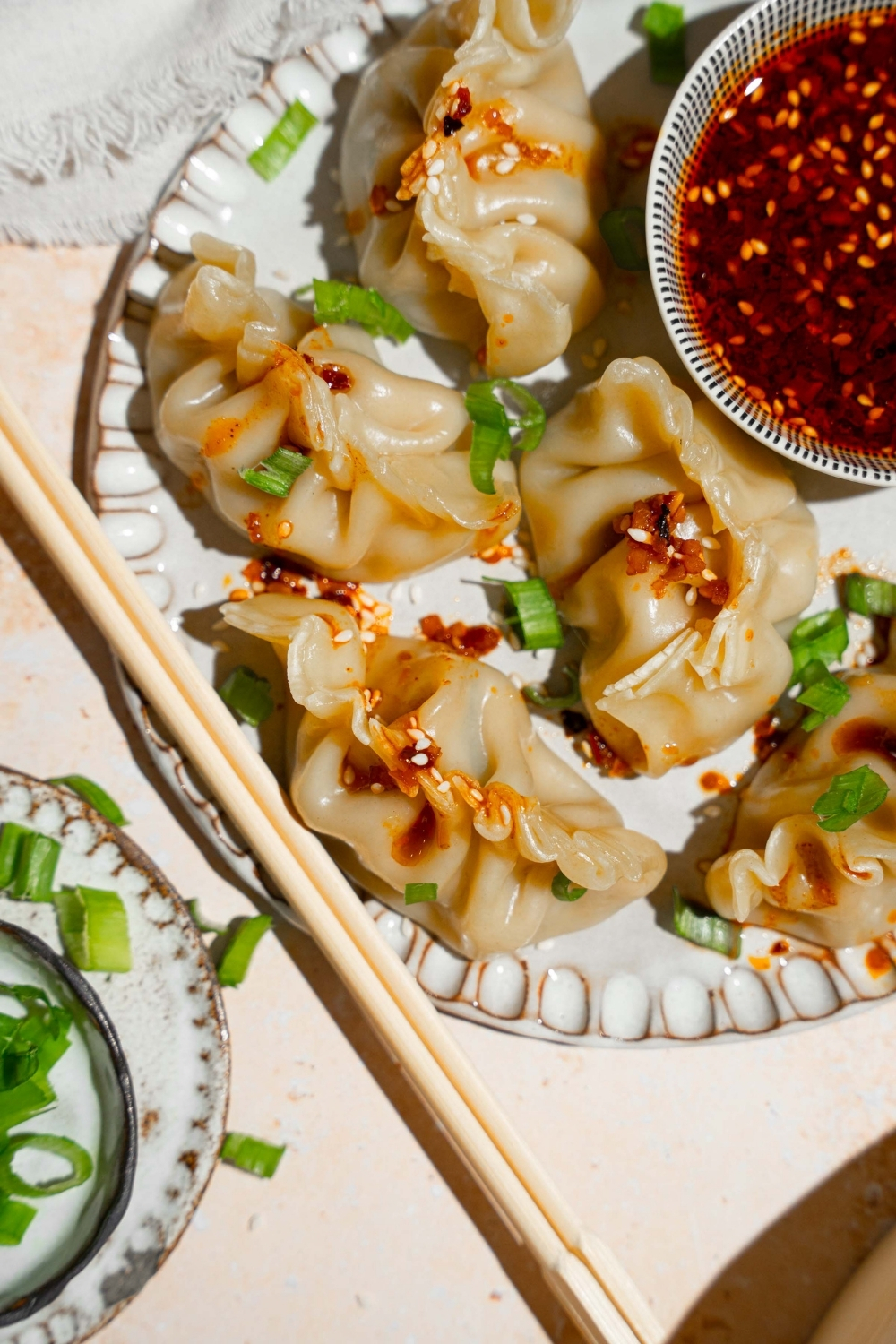 A plate with steamed chicken dumplings with a bowl of chili oil sauce. The plate is on a tan counter with a pair of chopsticks.