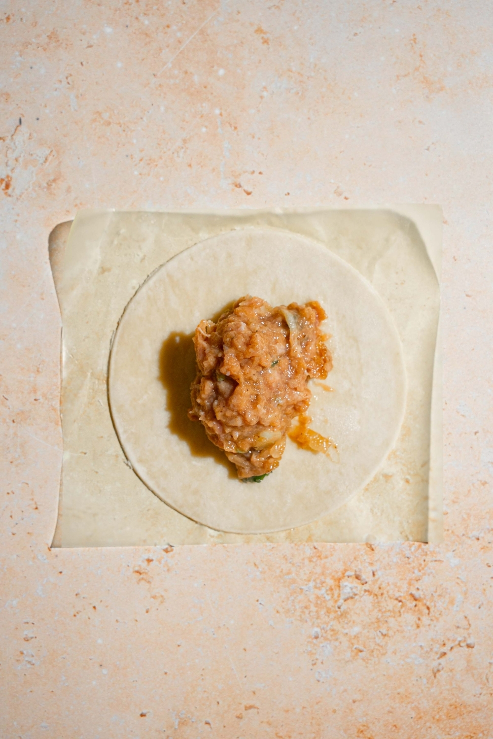 A piece of parchment paper with an open dumpling wrapper topped with a chicken mixture. The paper is on a tan counter.