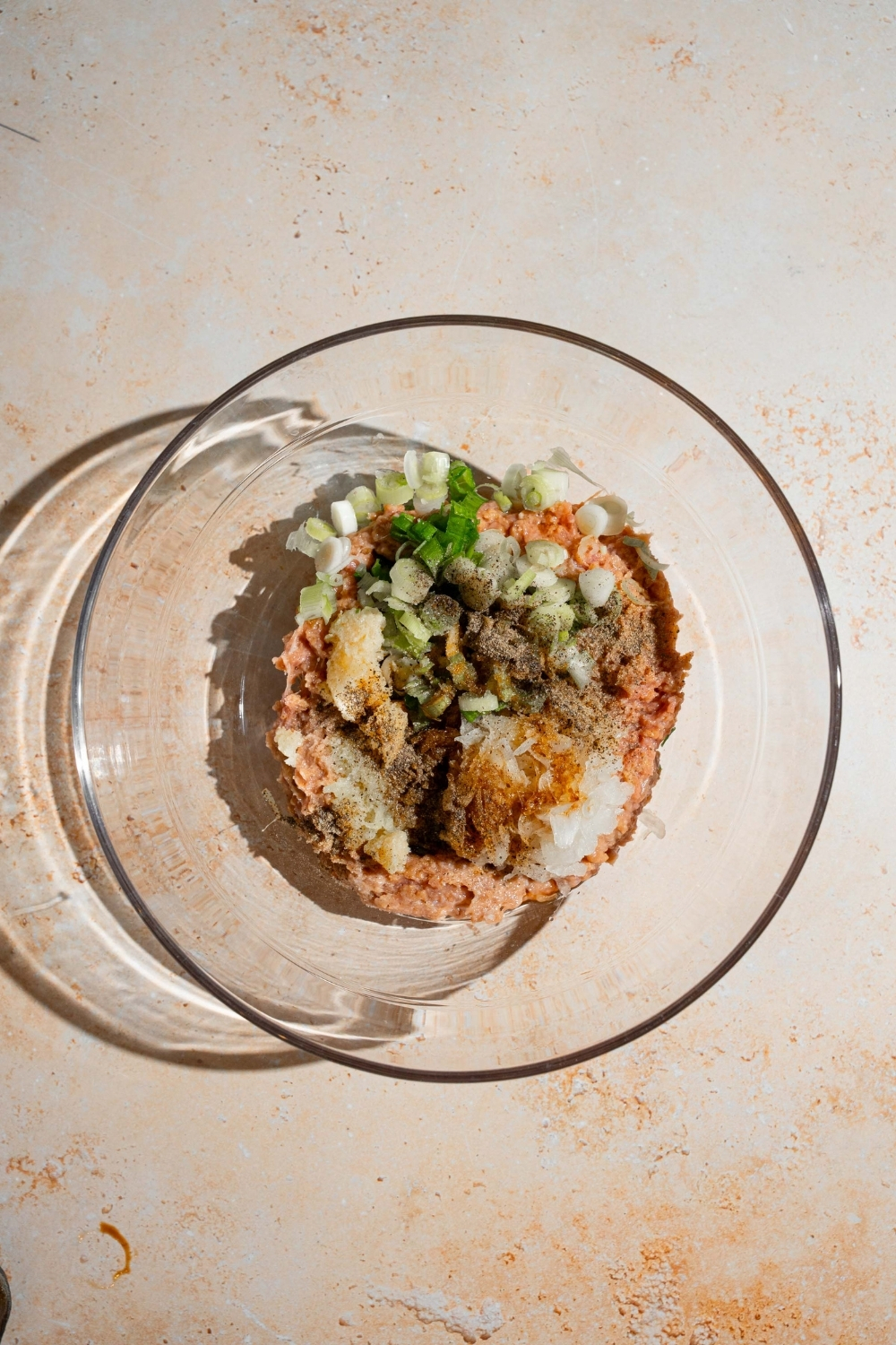 A glass bowl with ingredients to make chicken mixture for dumplings including chicken, green onion, ginger, garlic, soy sauce, and more. The bowl is on a tan counter.