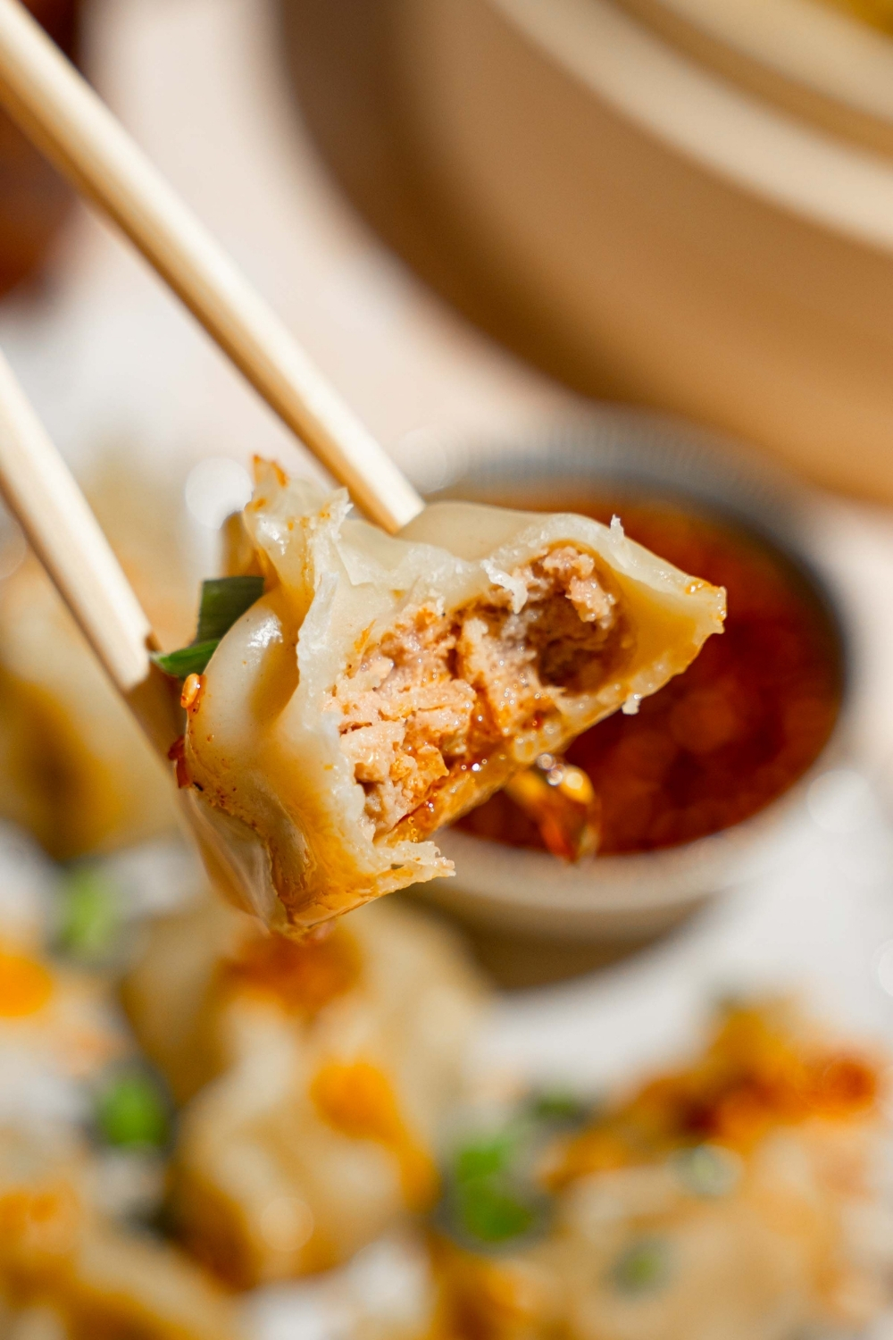 A close up of a pair of chopsticks with a steamed chicken dumpling with a bite taken from it. There is a plate of dumplings with a small bowl of chili oil sauce blurred in the background.